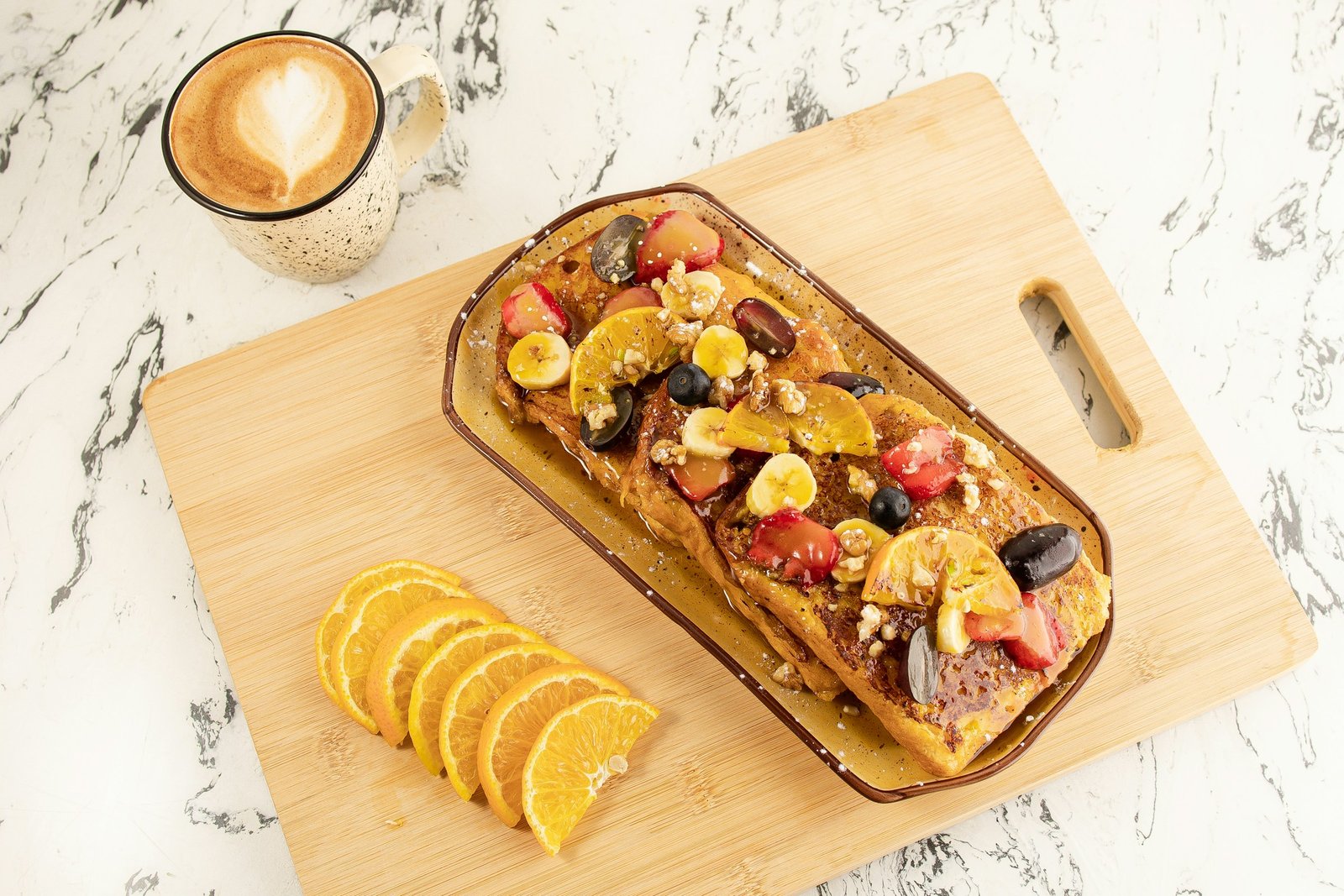 Wentelteefjes met verse fruitschijfjes en walnoten op een houten plank, naast een kop cappuccino en schijfjes sinaasappel