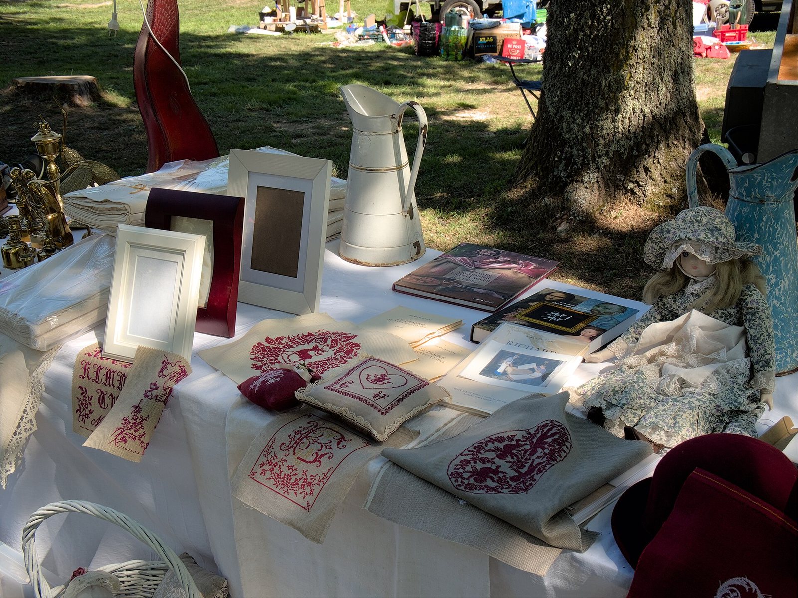 Tafel op een rommelmarkt met fotolijstjes, een witte lampetkan, borduurwerk en een pop, onder een boom op een grasveld vol kramen