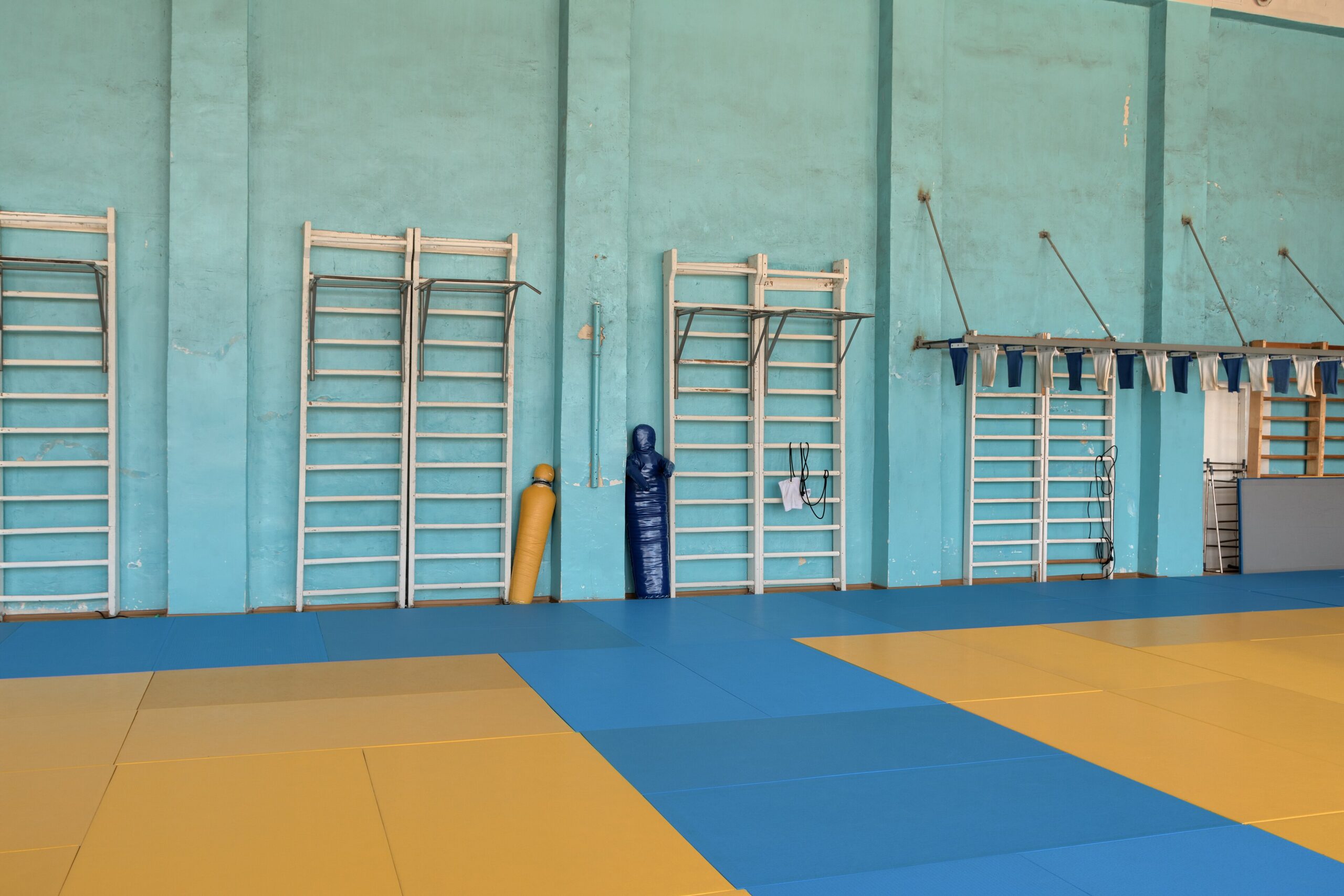 Lege sporthal met blauwe en gele valmatten op de vloer en houten rekken tegen een turquoise muur