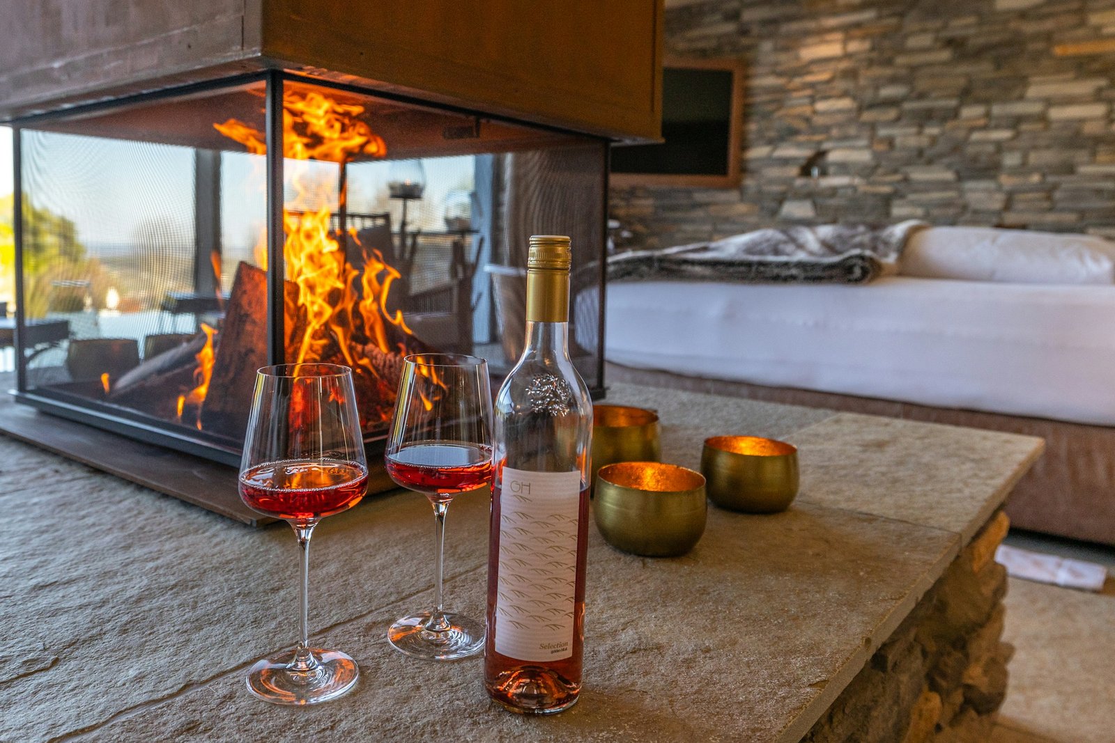 Twee glazen wijn en een fles wijn op een stenen tafel voor een brandende openhaard, met goudkleurige waxinelichtjes ernaast